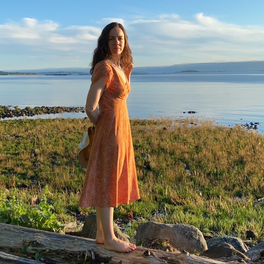 Me standing on a log on the beach with the water behind me, wearing a orange v-neck midi dress