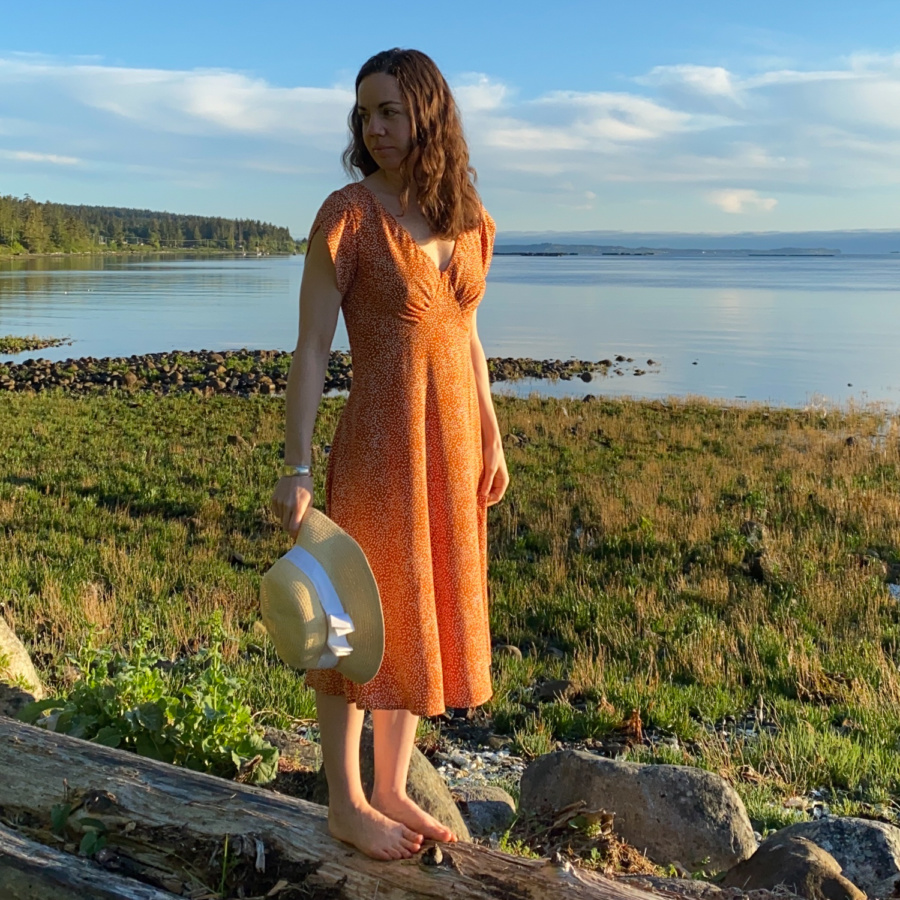 Me standing on a log on the beach with the water behind me, wearing a orange v-neck midi dress with white polka dots