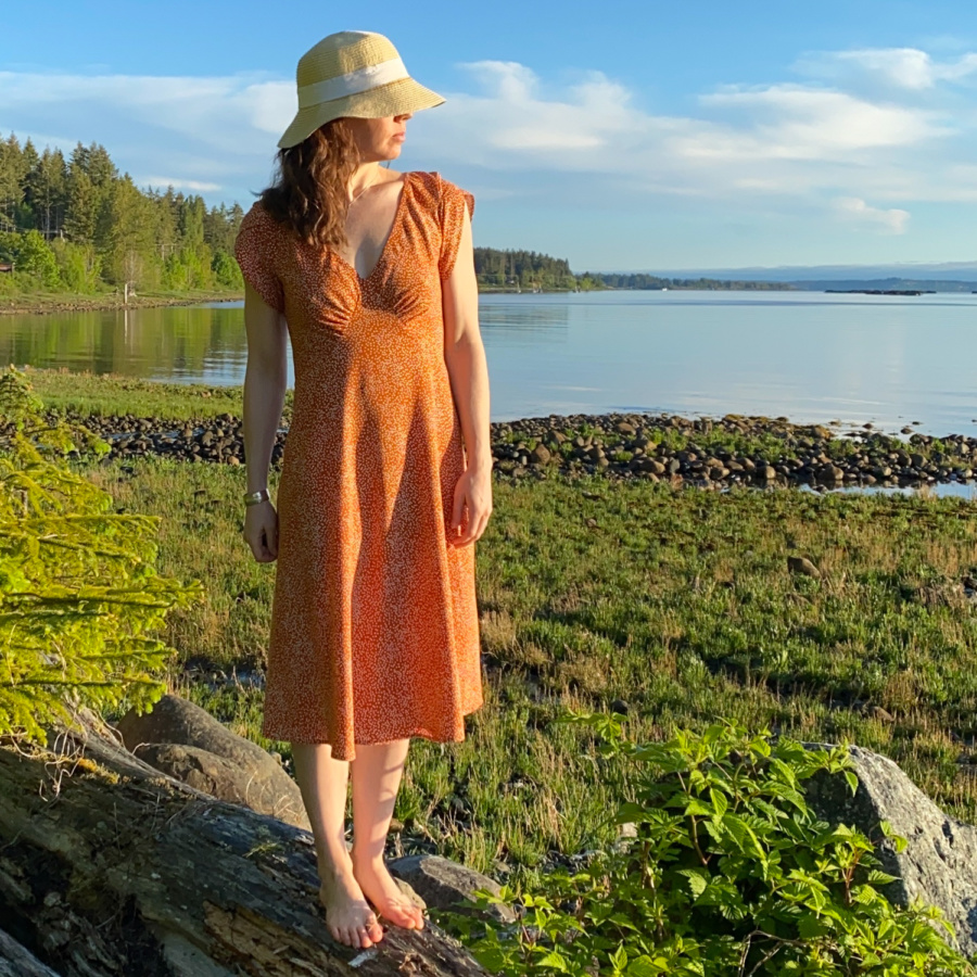 Me standing on a log on the beach with the water behind me, wearing a orange v-neck midi dress with white polka dots and a sun hat