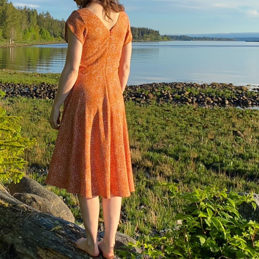 Me standing on a log on the beach with my back to the camera and the water behind me, wearing a orange v-back midi dress with white polka dots