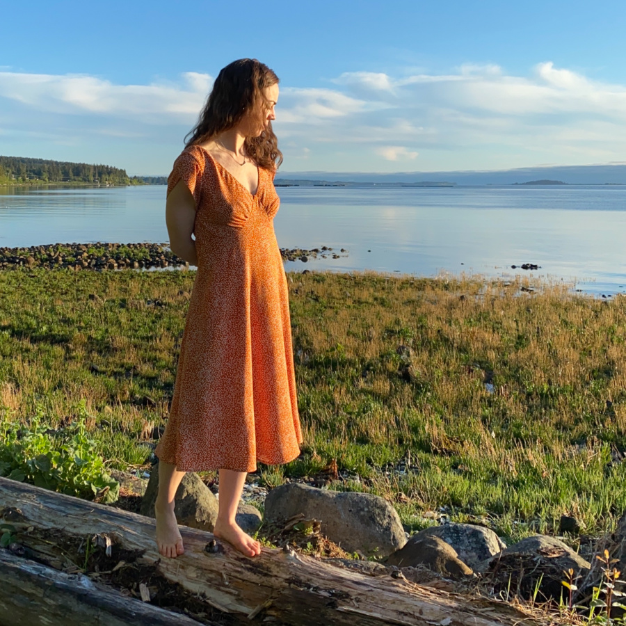 Me standing on a log on the beach with the water behind me, wearing a orange v-neck midi dress with white polka dots