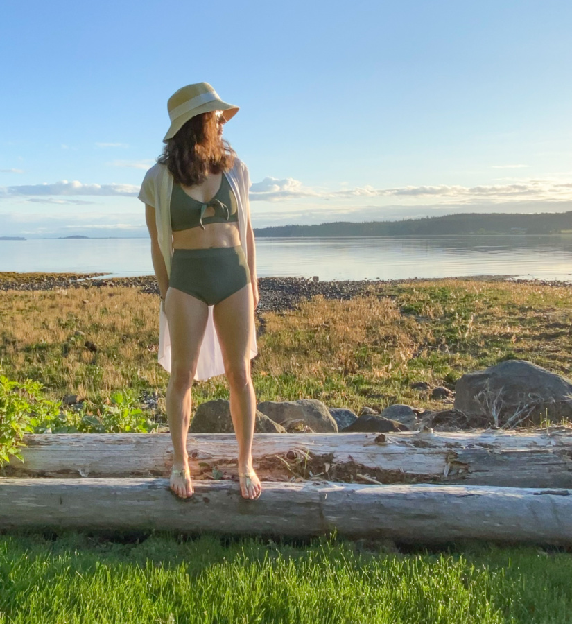Me standing on a log in front of a rocky beach wearing a sage green swim suit, white coverup and a hat