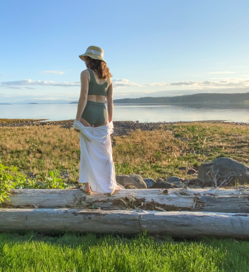 Me standing on a log in front of a rocky beach facing away from the camera wearing a sage green swim suit, white coverup and a hat