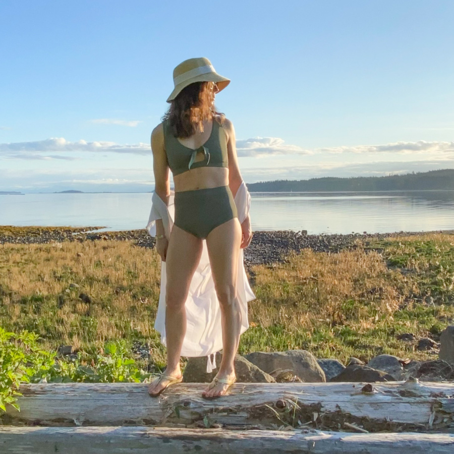 Me standing on a log in front of a rocky beach wearing a sage green swim suit and a hat