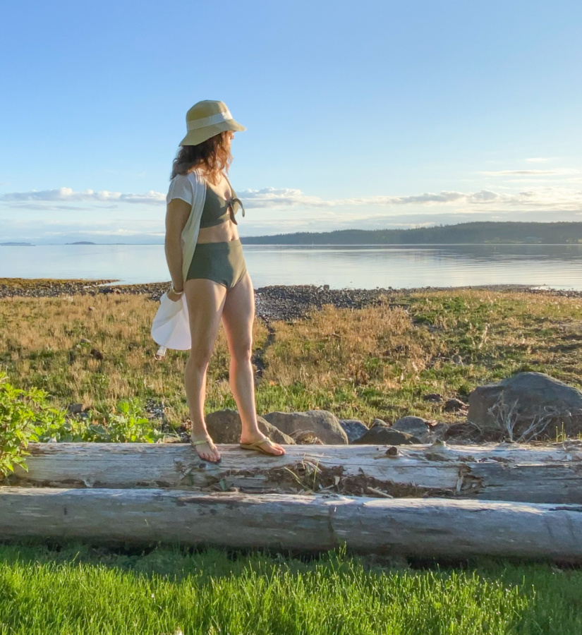 Me standing on a log in front of a rocky beach wearing a sage green swim suit and a hat