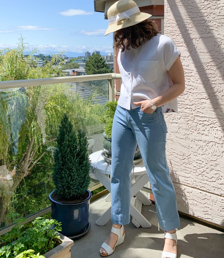 Me standing on a balcony wearing a cropped, white linen Kalle Shirt and blue jeans, looking away from the camera with one hand in my jeans pocket.