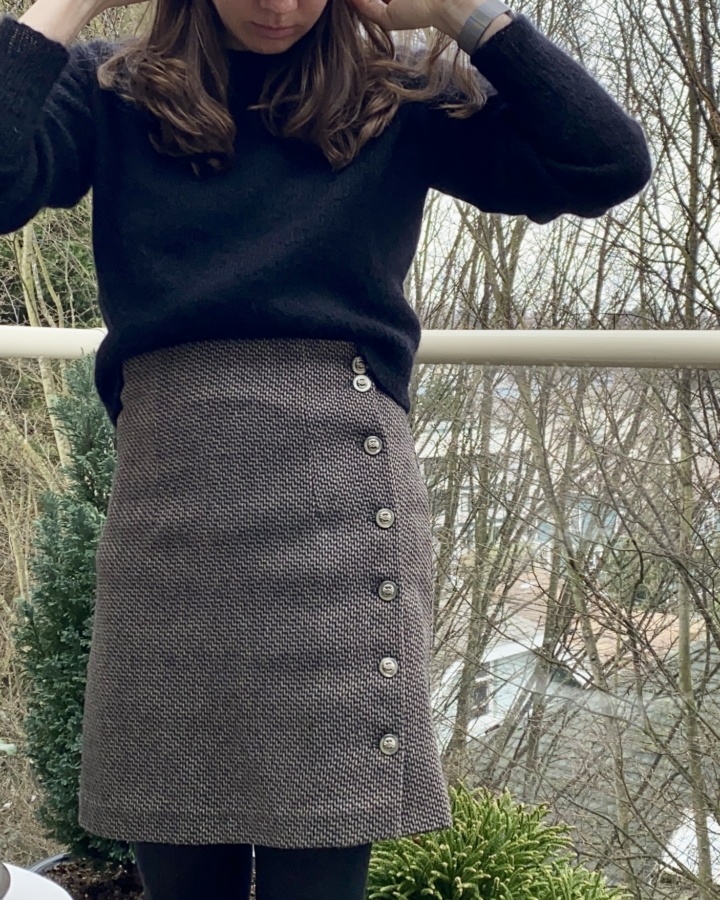 Close up front view of wool skirt with buttons running down the left side. Worn with a black fuzzy sweater tucked into the skirt at the front.