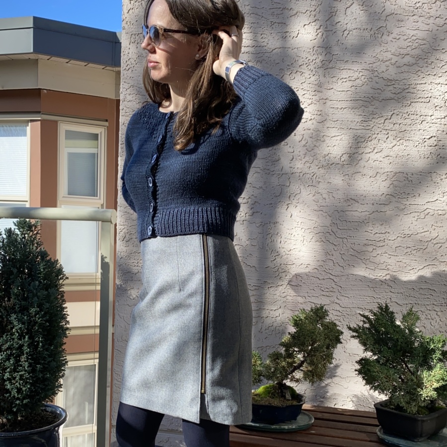 Standing in the sun, facing to my right, wearing a grey skirt with exposed metal zip at the side, navy blue buttoned cardigan and sunglasses. Shown from the knees up
