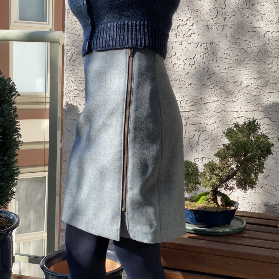 Close up side view of grey wool mini skirt with exposed metal zipper. Small bonsai tree behind.