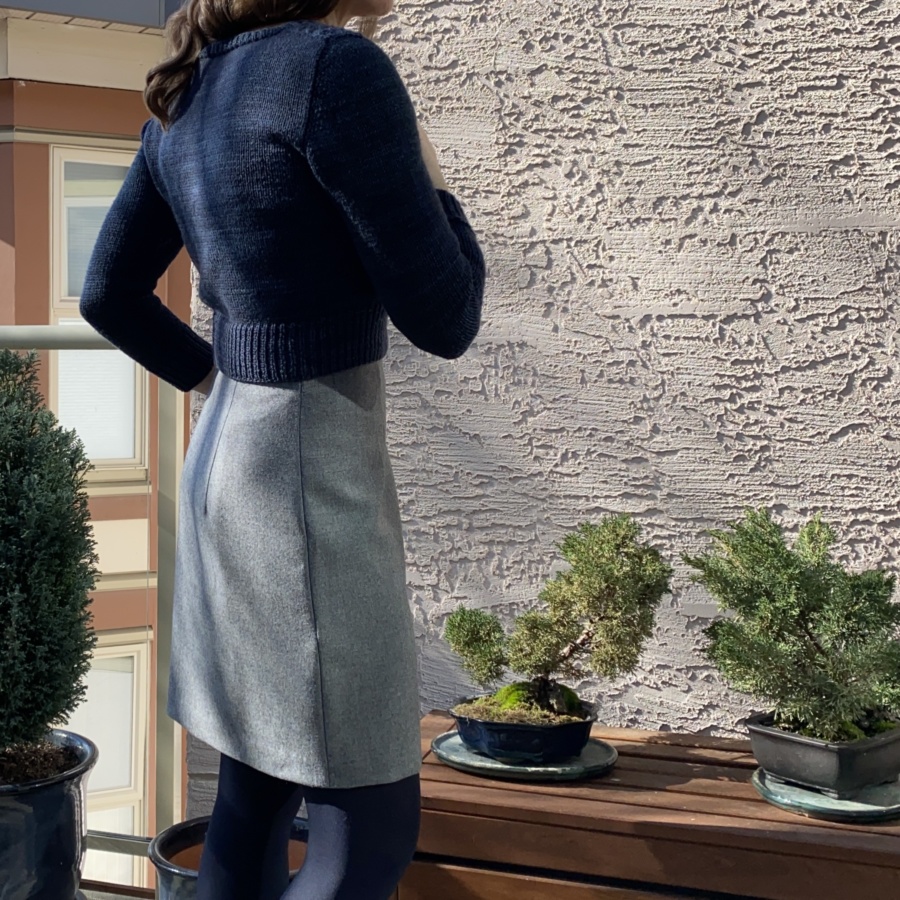 Right side/back view of me wearing navy blue sweater and grey mini skirt, shown from below shoulders to knees. Two small bonsai trees behind.