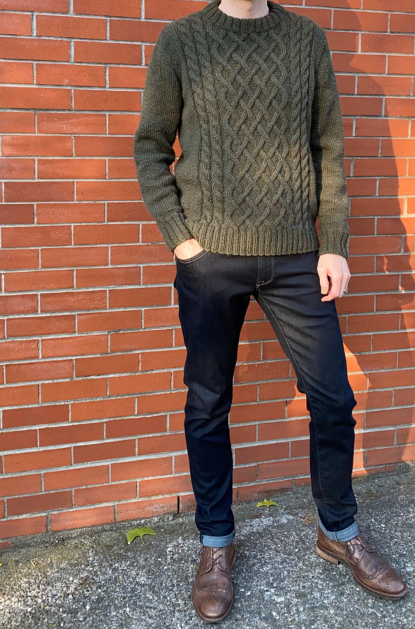 Tall, slim white man shown from the neck down wearing a forest-green cable knit sweater, dark blue jeans and brown lace up boots standing against a brick wall.