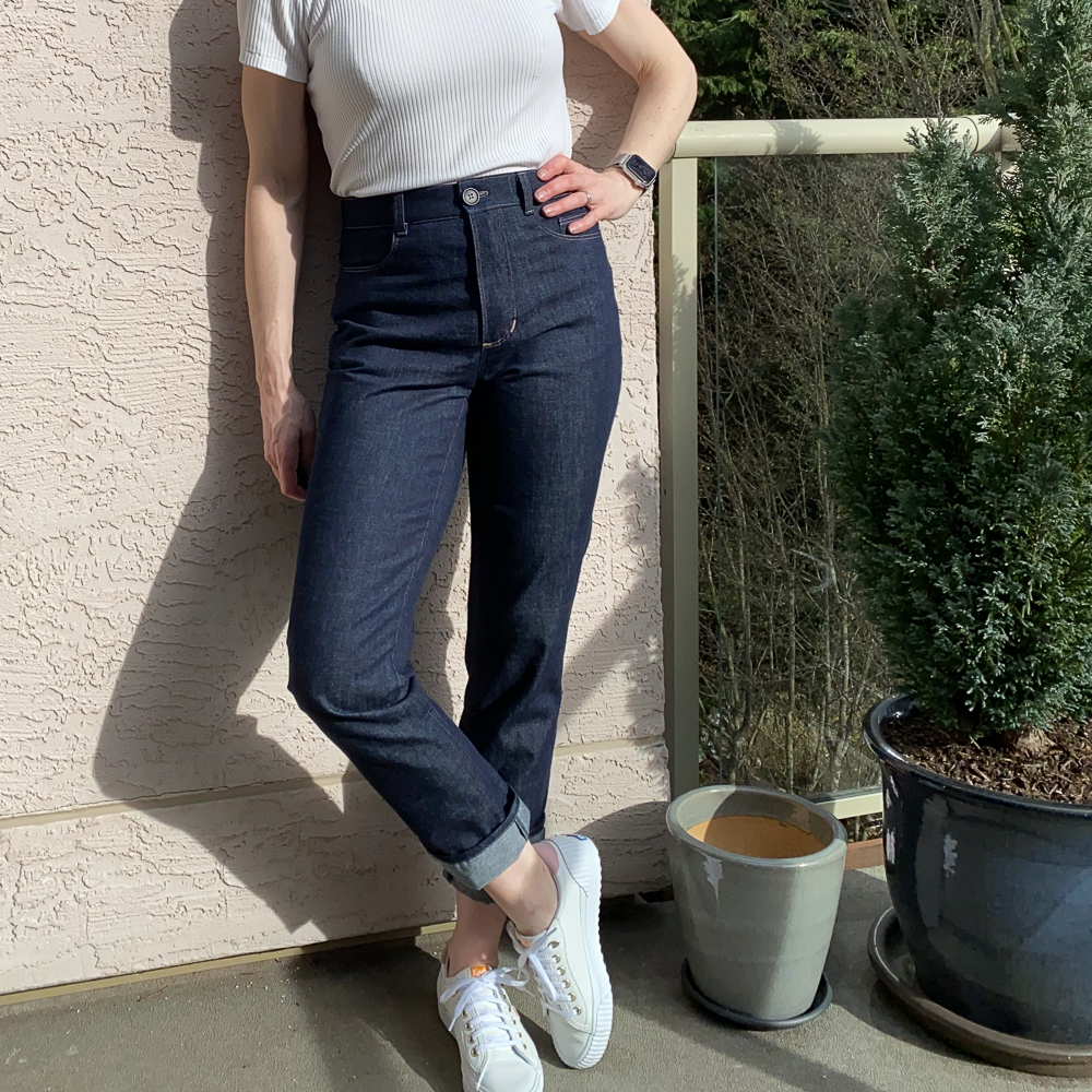 Me wearing my indigo jeans, leaning against a pink wall, beside a potted hedge tree. My left hand is on my hips. The photo is from the bust down. I'm wearing a white short sleeved turtleneck and white runners.