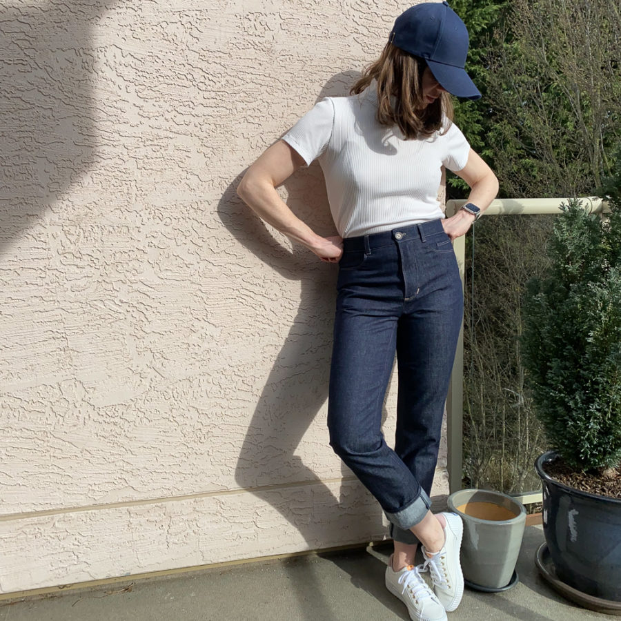 Me wearing my indigo jeans, leaning against a pink wall, beside a potted hedge tree. My hands are on my hips and I'm looking down. I'm wearing a white short sleeved turtleneck, white runners and a navy ball cap.