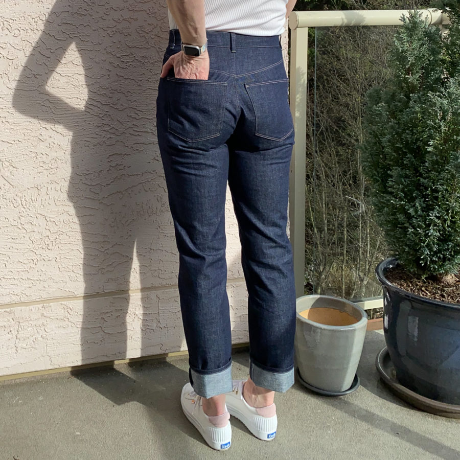 Me wearing my indigo jeans, standing beside a pink wall. Photo is from mid-back down and I'm facing away from the camera with my left hand in my left back pocket. I'm wearing a white top and white runners.