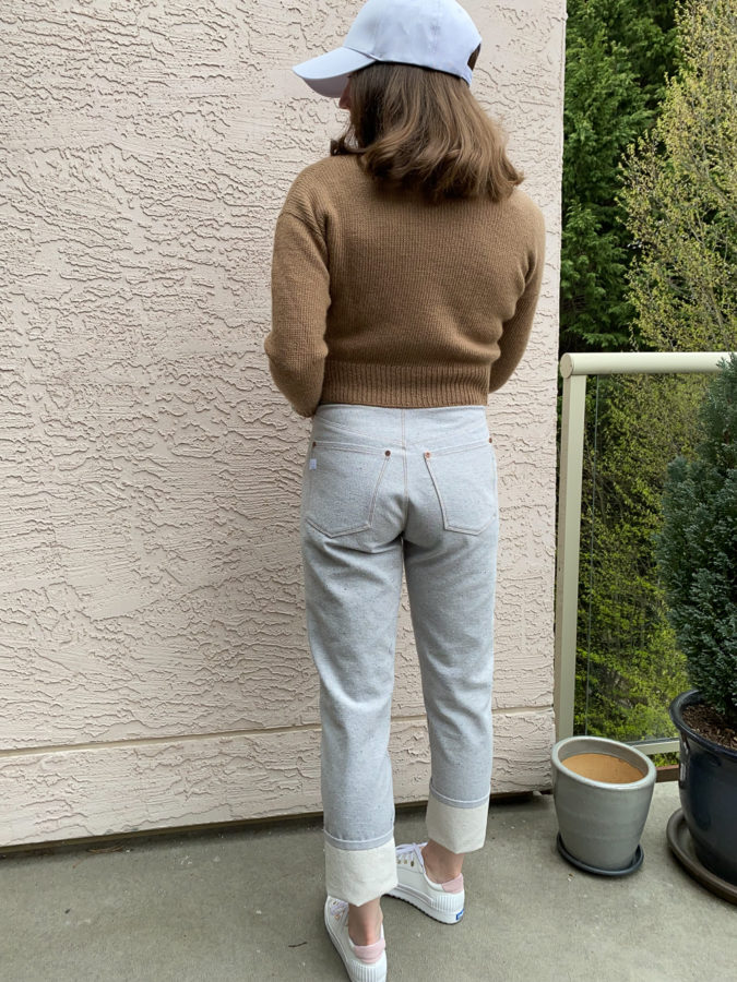 Me wearing my grey Brooks Jeans, standing against a pink wall. I'm facing away from the camera and am wearing a brown hand-knit sweater, white runners and a white ball cap.