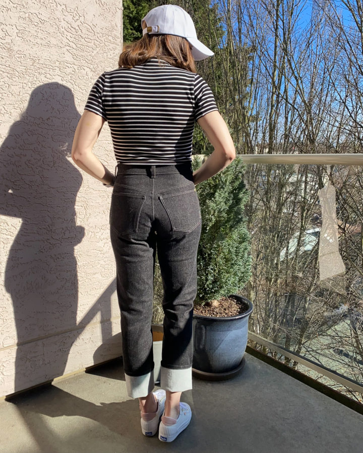 Me wearing my black Brooks Jeans, a black and white striped top, white runners and a white ball cap. I'm standing beside a small potted hedge tree and facing away from the camera.